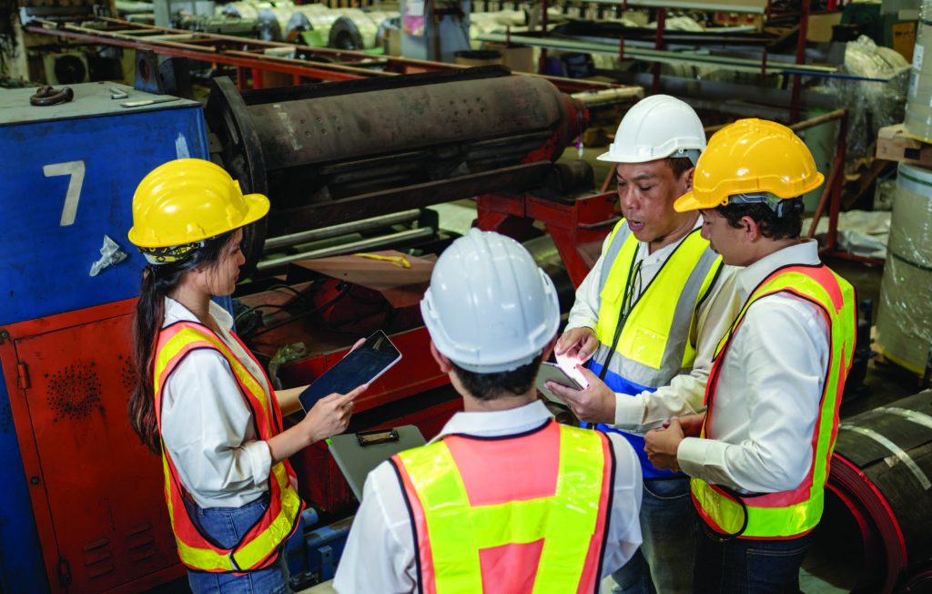 Four workers in safety helmets and vests reviewing a tablet in an industrial setting.